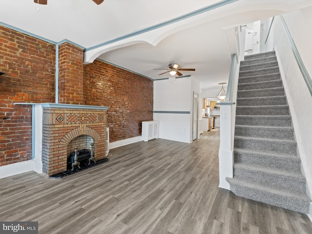 unfurnished living room featuring ceiling fan, wood-type flooring, a fireplace, and brick wall