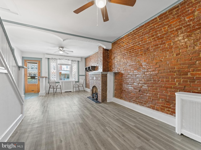 unfurnished living room with ceiling fan, brick wall, a fireplace, wood-type flooring, and radiator heating unit