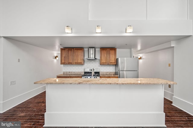 kitchen featuring an island with sink, stainless steel appliances, decorative backsplash, wall chimney exhaust hood, and light stone counters