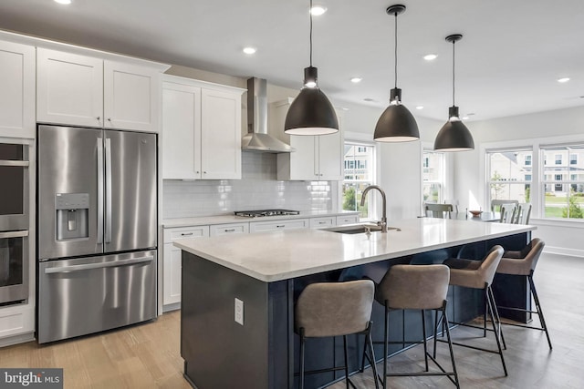 kitchen with wall chimney range hood, sink, appliances with stainless steel finishes, white cabinets, and a center island with sink