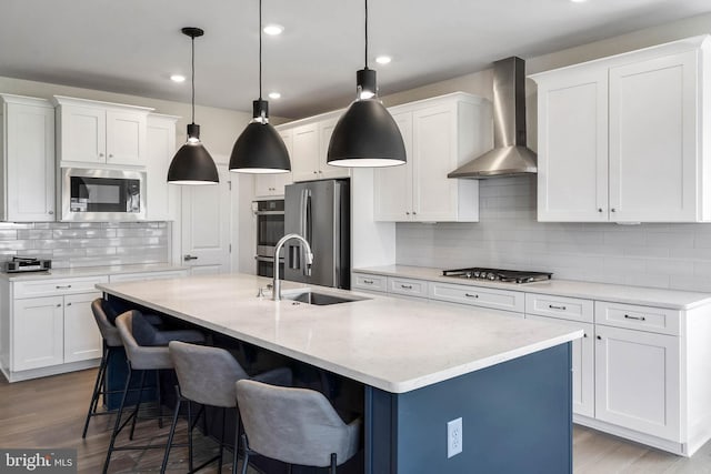 kitchen featuring a kitchen island with sink, hanging light fixtures, stainless steel appliances, and wall chimney exhaust hood