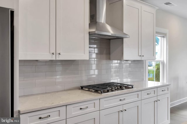 kitchen with stainless steel gas stovetop, white cabinets, backsplash, light stone counters, and wall chimney range hood