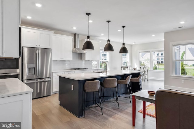 kitchen featuring an island with sink, white cabinetry, hanging light fixtures, stainless steel appliances, and wall chimney exhaust hood