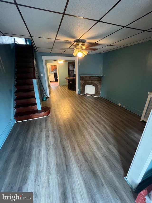 unfurnished living room with a drop ceiling, a brick fireplace, wood-type flooring, and ceiling fan