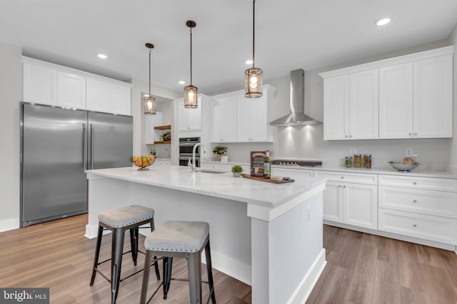 kitchen with pendant lighting, white cabinets, a kitchen island with sink, wall chimney range hood, and stainless steel appliances