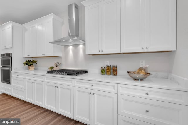 kitchen with wall chimney range hood, stainless steel appliances, white cabinets, and light hardwood / wood-style floors