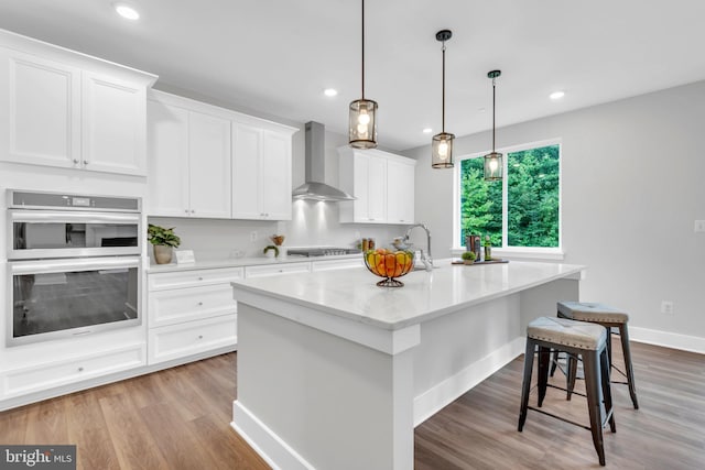 kitchen with stainless steel double oven, white cabinets, wall chimney exhaust hood, decorative light fixtures, and a kitchen island with sink