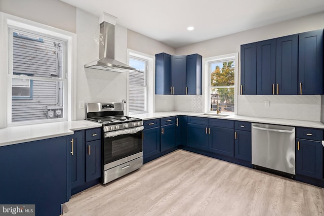 kitchen with blue cabinetry, appliances with stainless steel finishes, wall chimney range hood, and a sink