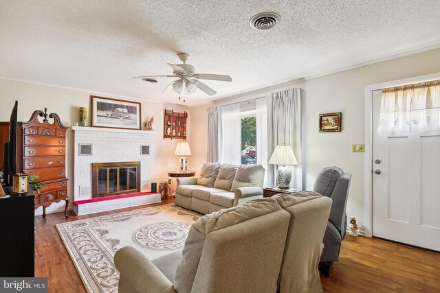 living room featuring crown molding, a textured ceiling, a brick fireplace, ceiling fan, and wood-type flooring