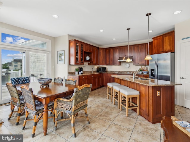 kitchen featuring decorative light fixtures, sink, light stone counters, stainless steel refrigerator with ice dispenser, and a center island with sink