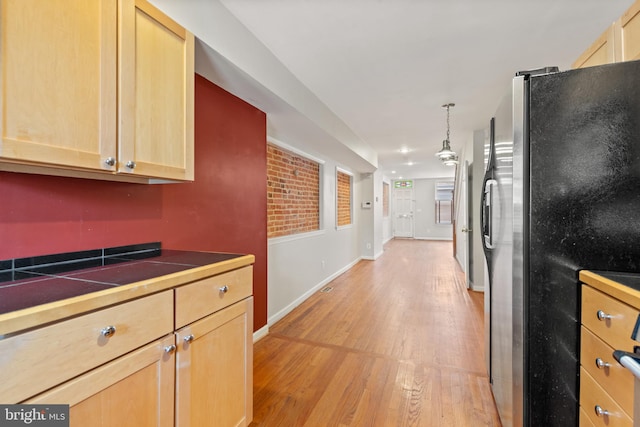 kitchen with tile countertops, light brown cabinetry, stainless steel fridge, hanging light fixtures, and light hardwood / wood-style floors