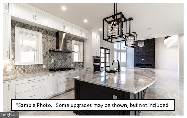 kitchen with wall chimney range hood, stainless steel appliances, white cabinets, sink, and a kitchen island with sink