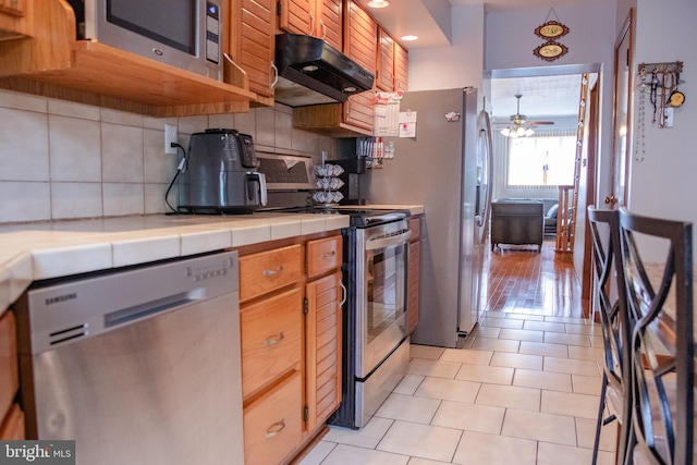 kitchen with stainless steel appliances, light hardwood / wood-style floors, decorative backsplash, tile counters, and ceiling fan