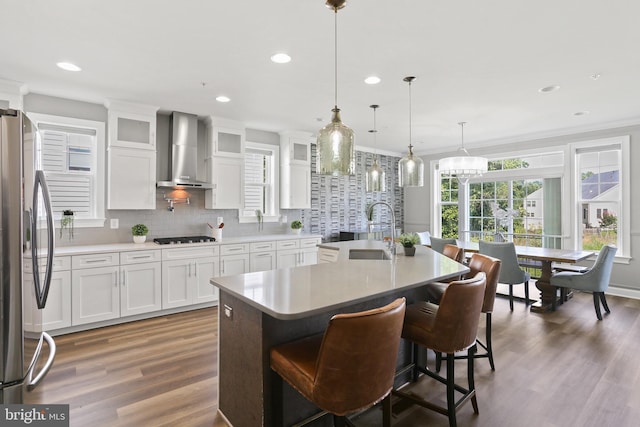 kitchen with glass insert cabinets, freestanding refrigerator, a sink, an island with sink, and wall chimney exhaust hood