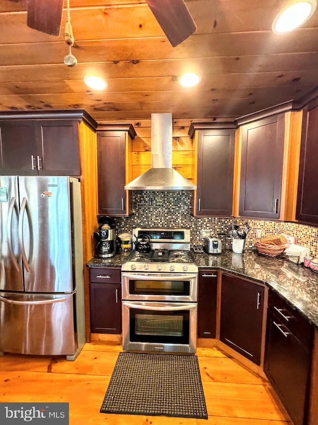 kitchen featuring tasteful backsplash, light wood-type flooring, dark stone counters, wall chimney exhaust hood, and appliances with stainless steel finishes