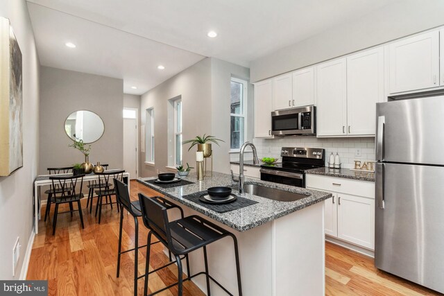 kitchen with dark stone countertops, a kitchen island with sink, appliances with stainless steel finishes, tasteful backsplash, and light hardwood / wood-style floors