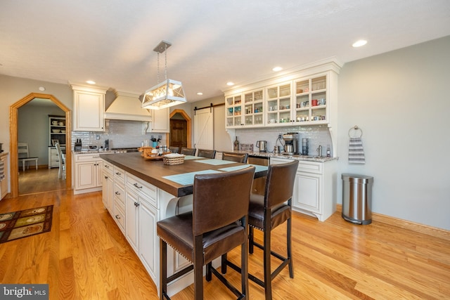 kitchen with butcher block counters, decorative backsplash, a barn door, custom range hood, and light hardwood / wood-style flooring