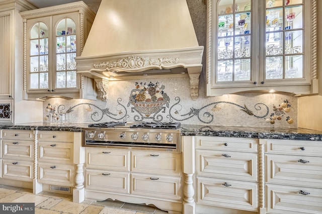 kitchen with backsplash, stainless steel gas stovetop, custom exhaust hood, cream cabinetry, and dark stone counters