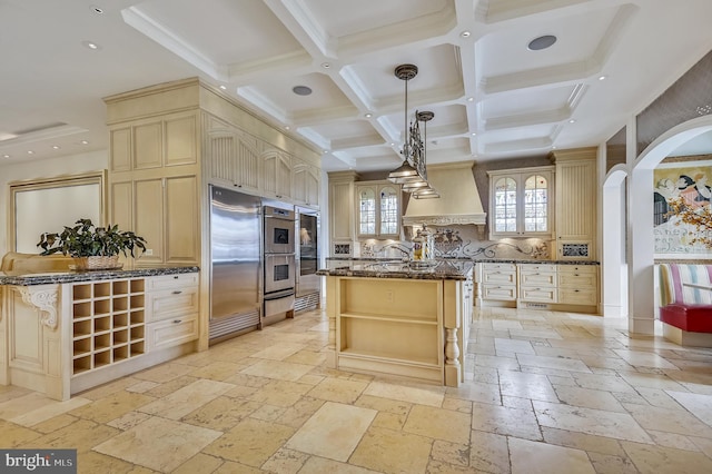 kitchen featuring premium range hood, built in refrigerator, decorative light fixtures, dark stone countertops, and a kitchen island