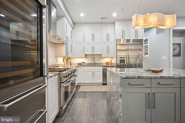 kitchen featuring a kitchen island, white cabinets, premium appliances, hanging light fixtures, and wall chimney range hood