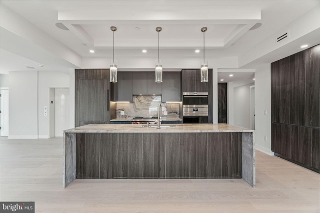 kitchen featuring sink, a kitchen island with sink, double oven, a tray ceiling, and decorative light fixtures