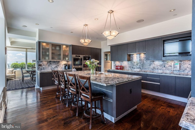 kitchen featuring hanging light fixtures, stainless steel double oven, a center island with sink, and dark hardwood / wood-style floors