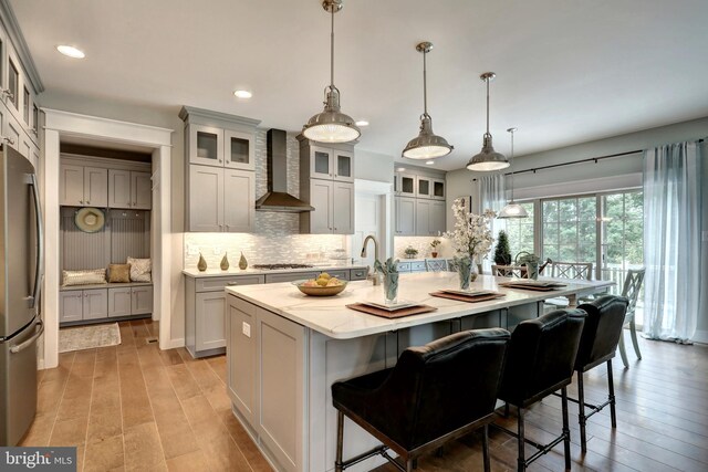 kitchen featuring an island with sink, appliances with stainless steel finishes, decorative light fixtures, and wall chimney exhaust hood