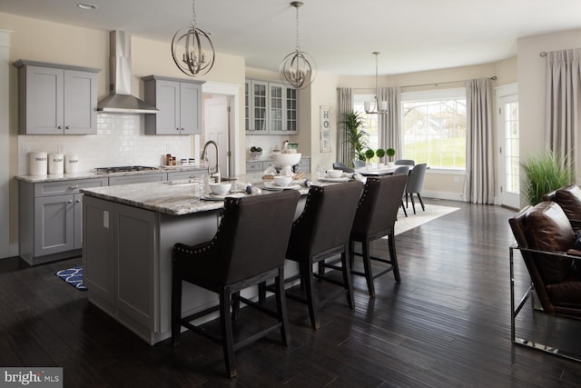 kitchen with wall chimney range hood, gray cabinetry, a kitchen island with sink, stainless steel gas cooktop, and a kitchen bar