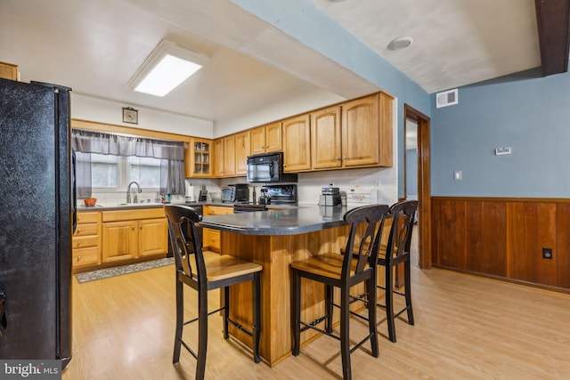 kitchen with a wainscoted wall, a breakfast bar area, visible vents, glass insert cabinets, and black appliances