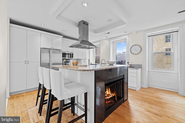 kitchen with extractor fan, light wood-type flooring, a kitchen island, a raised ceiling, and white cabinets