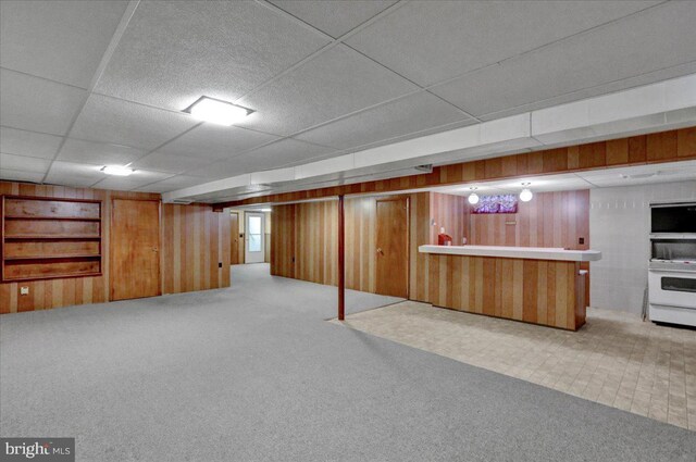 basement featuring light carpet, built in features, wooden walls, and a paneled ceiling