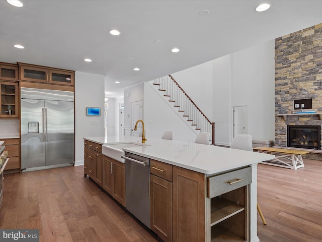 kitchen with a stone fireplace, an island with sink, wood-type flooring, sink, and stainless steel appliances