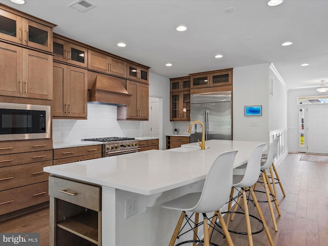 kitchen featuring a large island with sink, premium range hood, built in appliances, and a kitchen breakfast bar