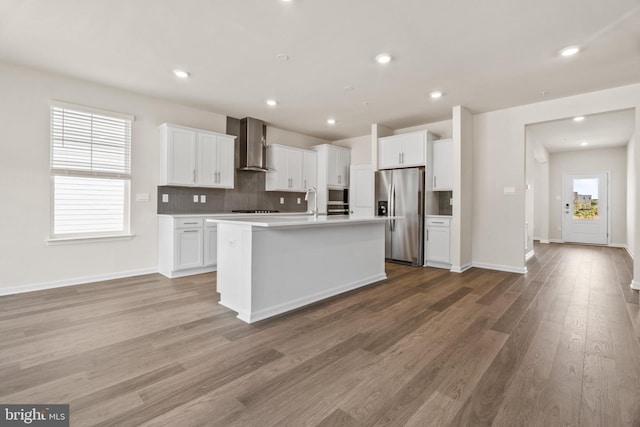 kitchen featuring wall chimney range hood, appliances with stainless steel finishes, white cabinets, a center island with sink, and decorative backsplash