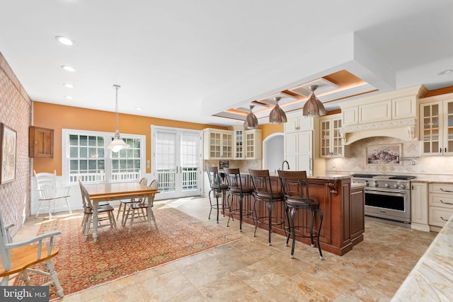 kitchen with high end stove, cream cabinets, hanging light fixtures, and a center island with sink