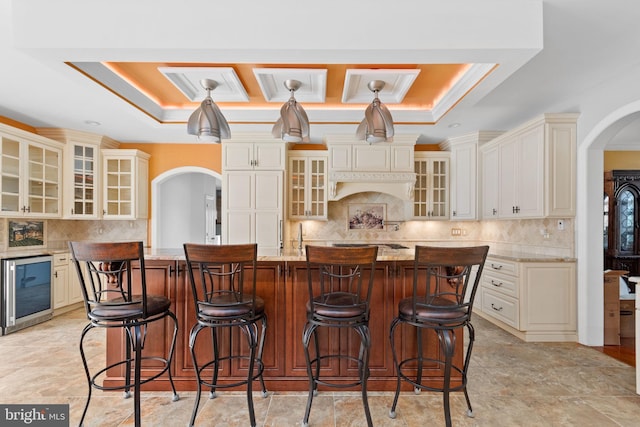 kitchen featuring a tray ceiling, light stone countertops, cream cabinets, and a center island with sink