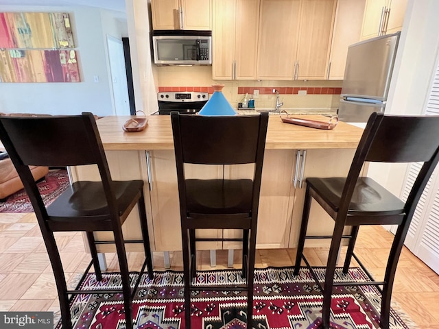 kitchen with light brown cabinetry, appliances with stainless steel finishes, a kitchen breakfast bar, decorative backsplash, and butcher block counters