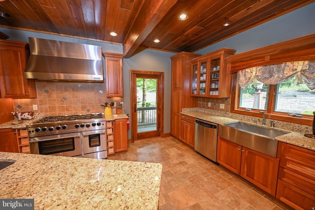 kitchen featuring appliances with stainless steel finishes, glass insert cabinets, a sink, wall chimney range hood, and light stone countertops