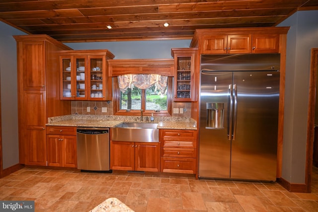 kitchen with light stone counters, a sink, appliances with stainless steel finishes, tasteful backsplash, and glass insert cabinets