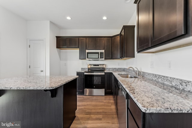 kitchen featuring stainless steel appliances, wood finished floors, a sink, dark brown cabinets, and light stone countertops