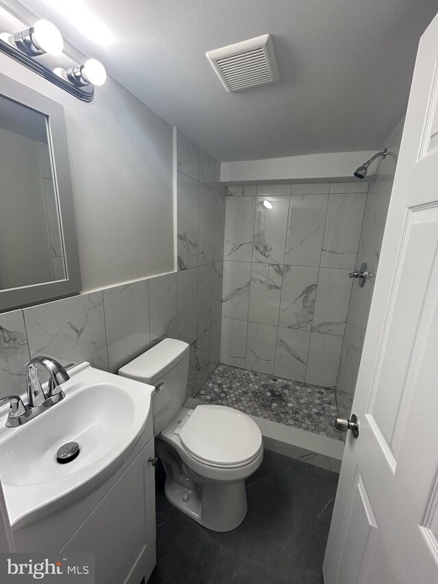 bathroom featuring tile patterned flooring, tiled shower, toilet, vanity, and tile walls