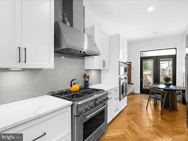 kitchen featuring white cabinetry, wall chimney range hood, stainless steel appliances, light parquet flooring, and light stone countertops