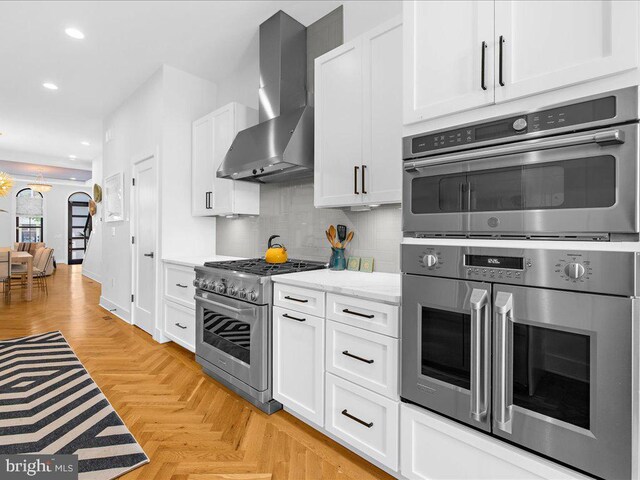 kitchen with white cabinetry, wall chimney range hood, light parquet floors, light stone counters, and appliances with stainless steel finishes