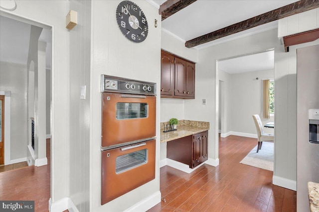 kitchen with light stone counters, multiple ovens, hardwood / wood-style flooring, dark brown cabinetry, and beamed ceiling