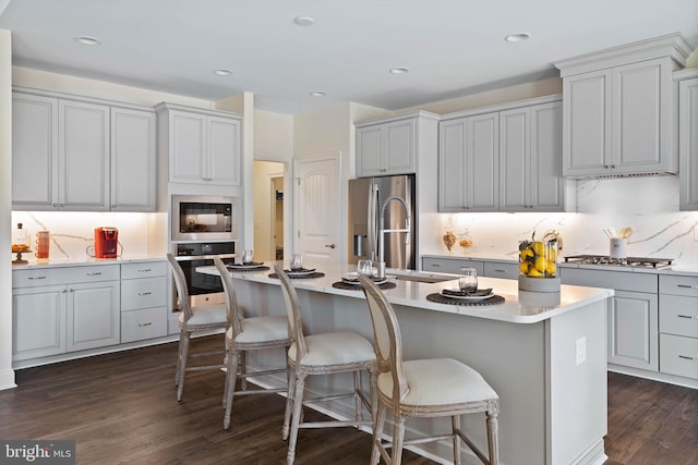 kitchen featuring dark hardwood / wood-style flooring, gray cabinetry, a kitchen island with sink, backsplash, and appliances with stainless steel finishes