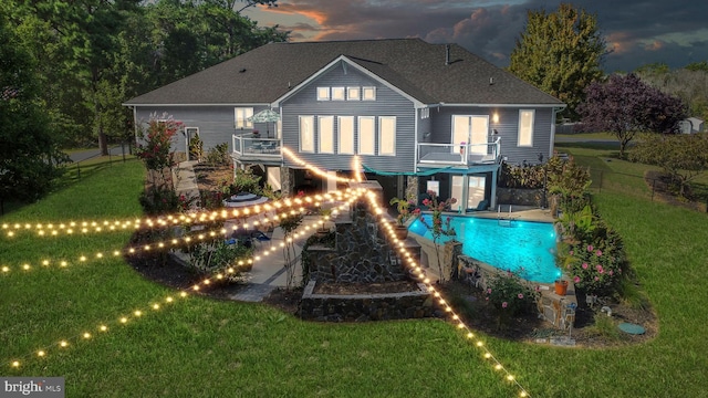 back house at dusk featuring a balcony, a fenced in pool, and a lawn