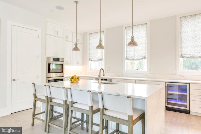 kitchen with stainless steel double oven, a kitchen island with sink, beverage cooler, light hardwood / wood-style floors, and sink