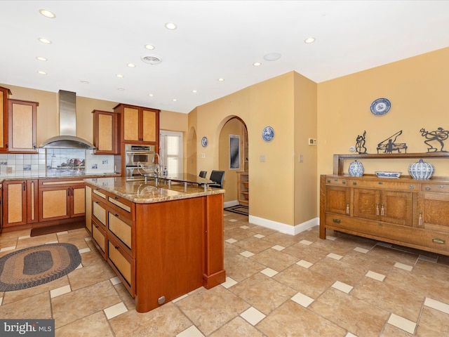 kitchen featuring light stone counters, a center island with sink, stainless steel double oven, wall chimney range hood, and backsplash