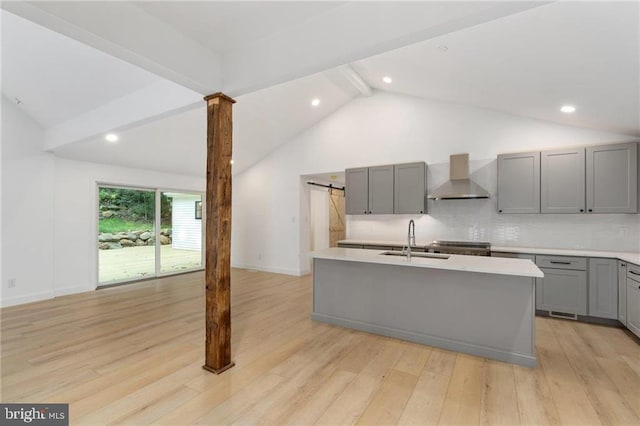 kitchen with light hardwood / wood-style flooring, wall chimney range hood, sink, beam ceiling, and an island with sink