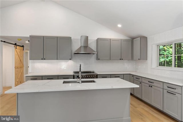 kitchen featuring lofted ceiling, wall chimney range hood, light stone counters, a barn door, and light hardwood / wood-style floors